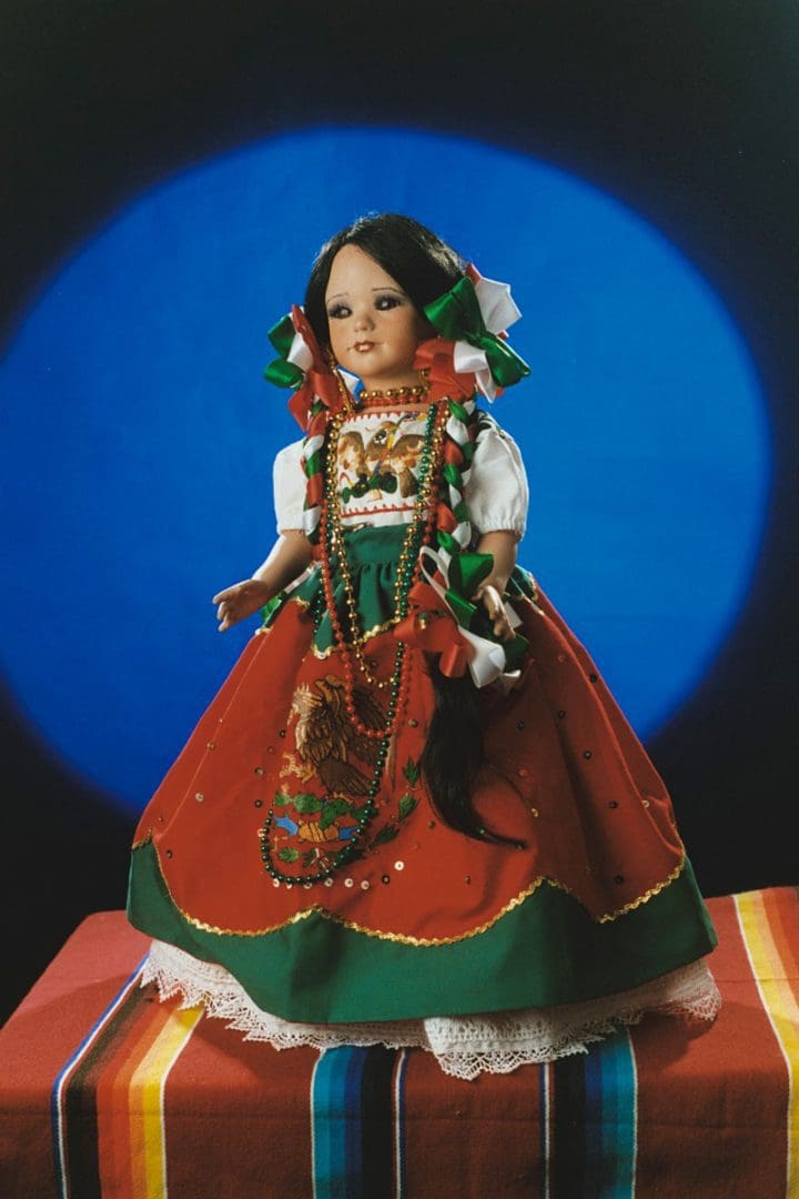 A doll wears a red, green, and white dress decorated with the Mexican eagle. She also wears red, green, and white ribbons in her long, dark hair
