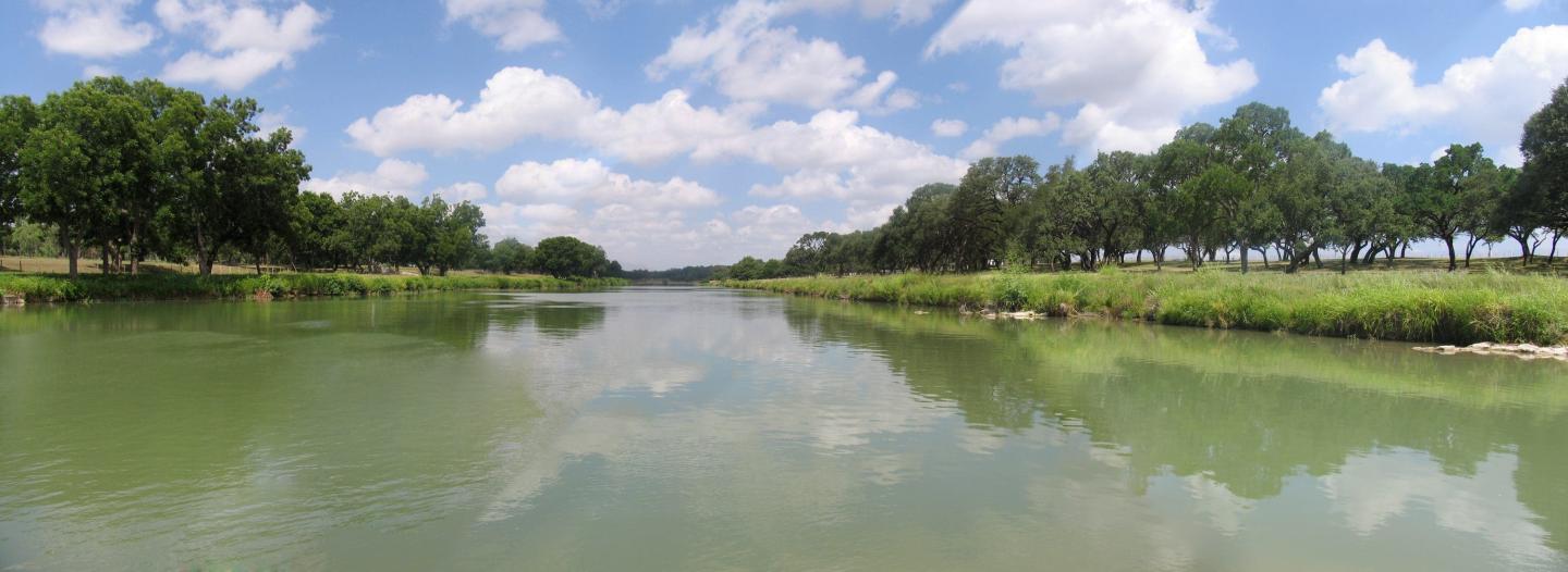 Calm river flanked by trees under a blue sky with clouds.
