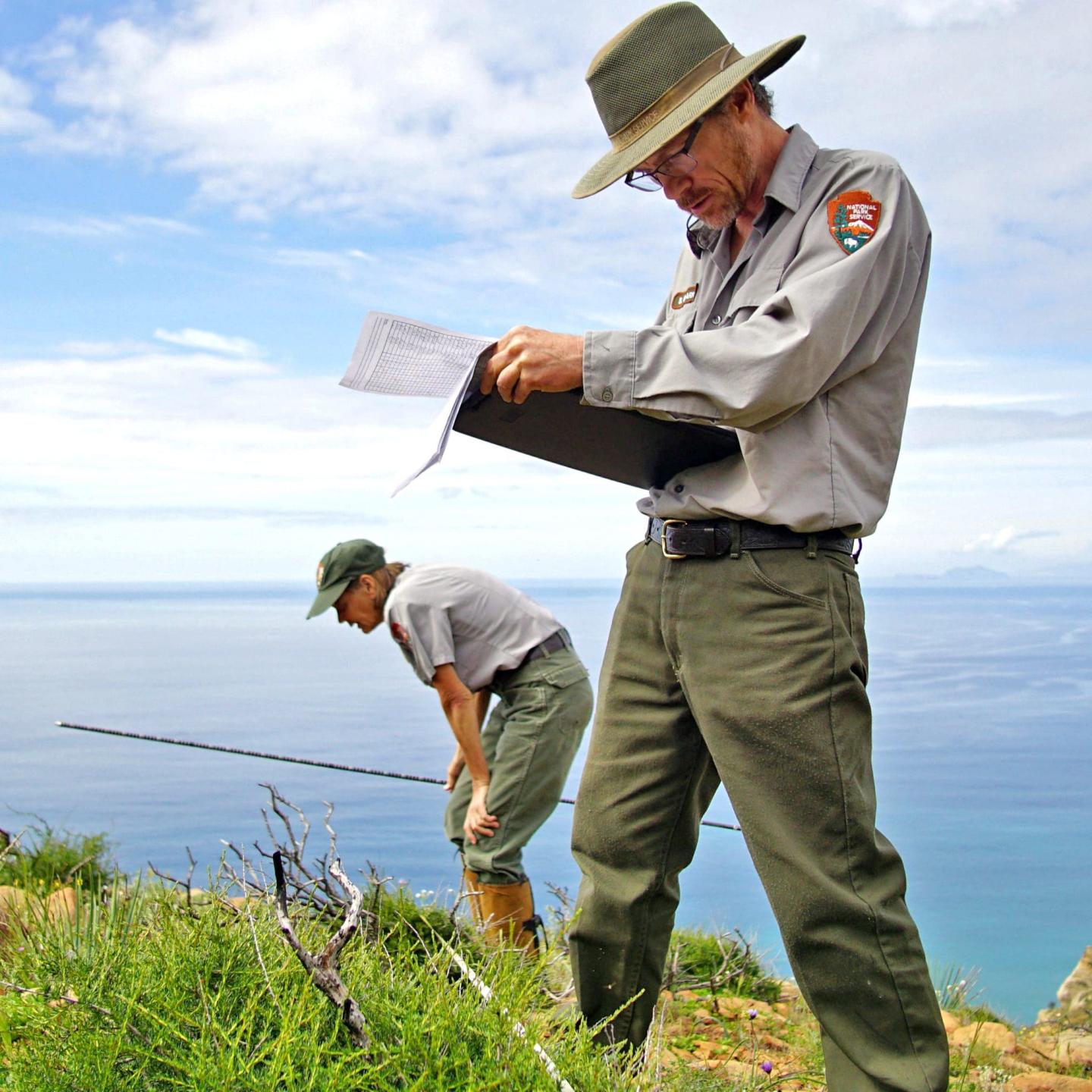 Two park rangers on a cliffside, one reading a clipboard, overlooking the ocean.