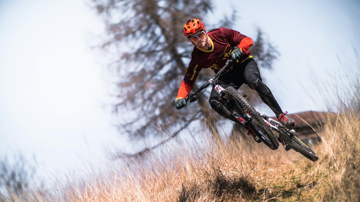 Mountain biker in red, riding downhill through tall grass.