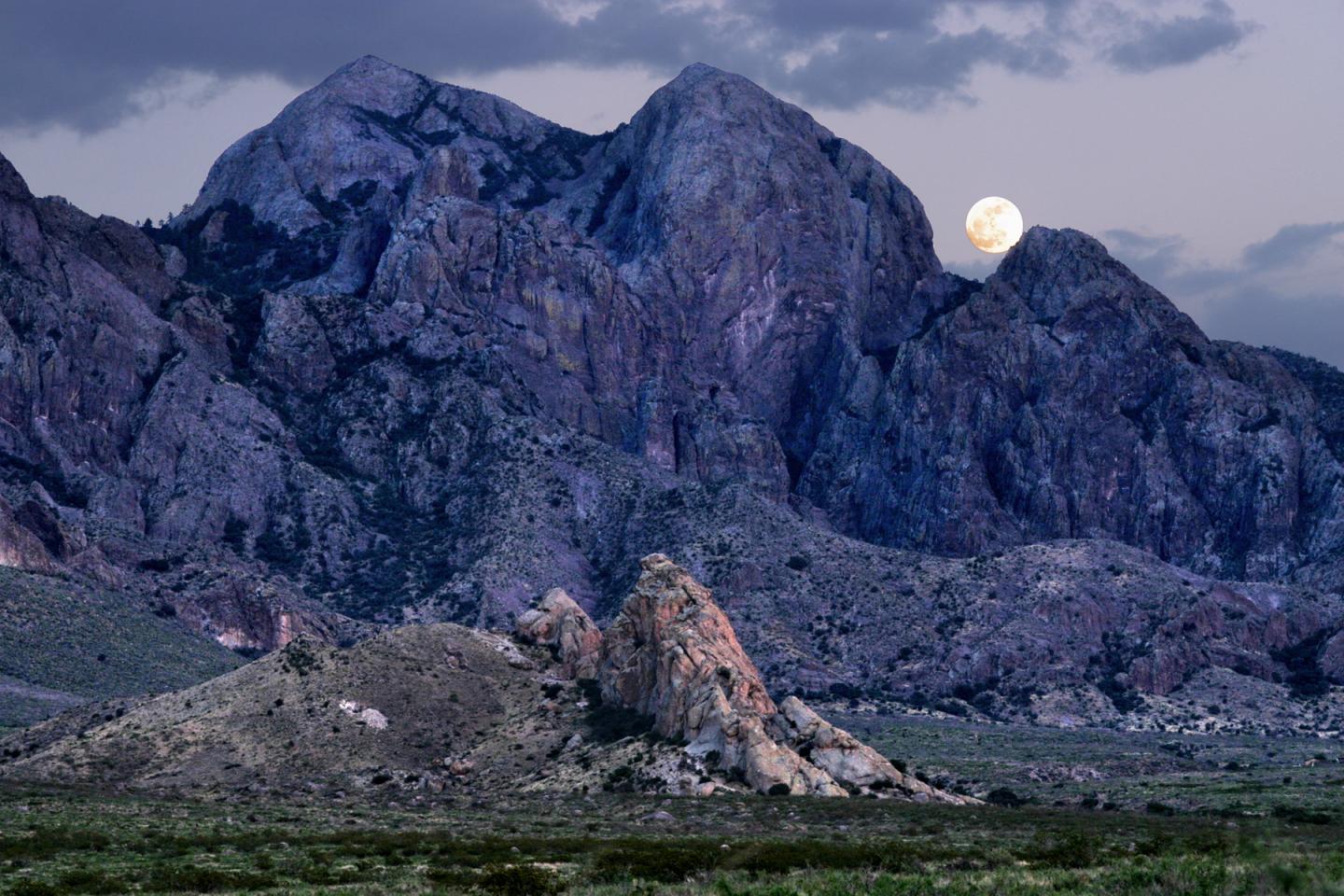 Desert mountain landscape at dusk with a full moon rising behind rocky peaks.