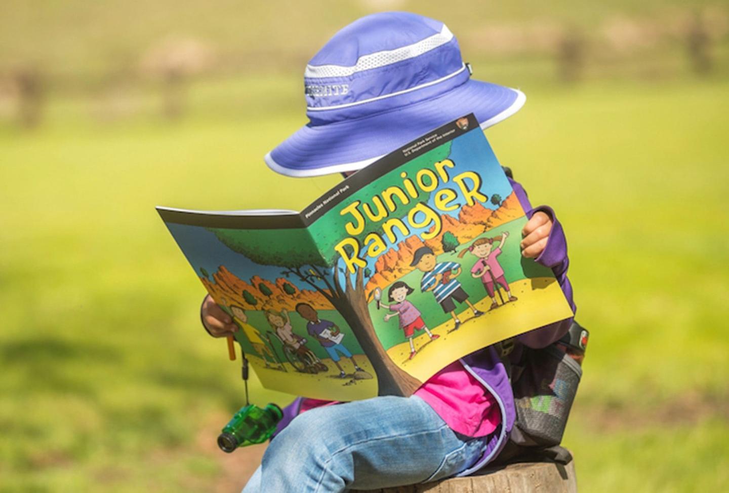 Child in a purple hat reading "Junior Ranger" in a grassy field.