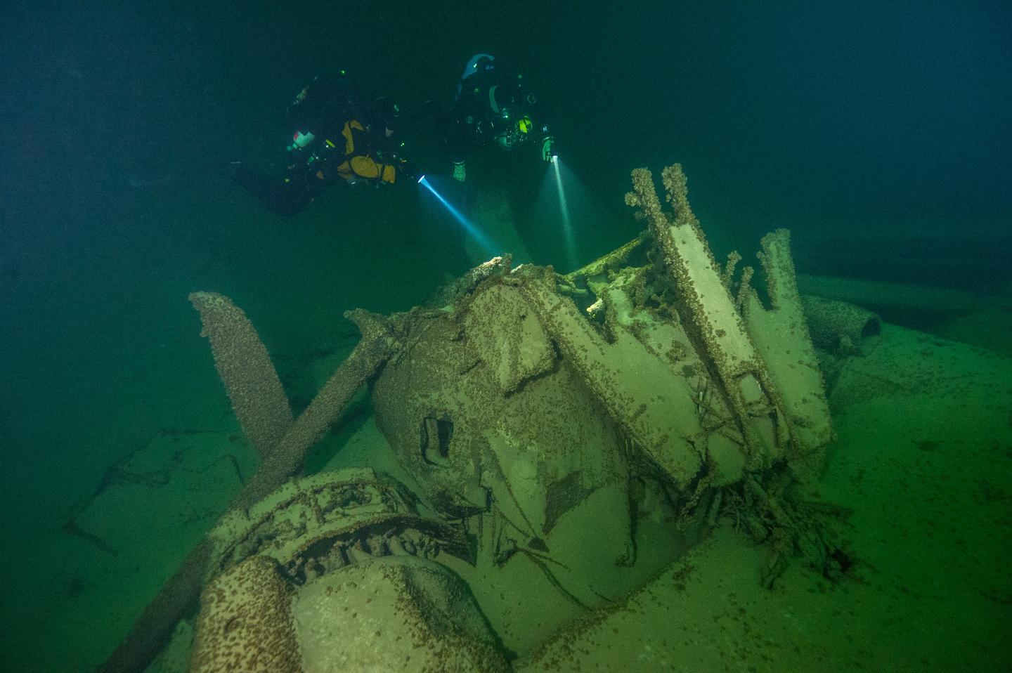 Divers explore a sunken aircraft underwater, using flashlights in dim lighting.