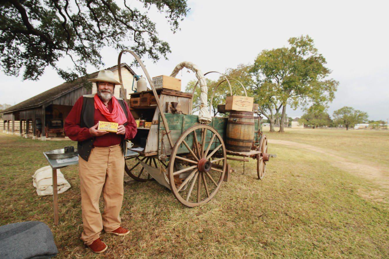 Cowboy standing beside a chuckwagon in a grassy field.