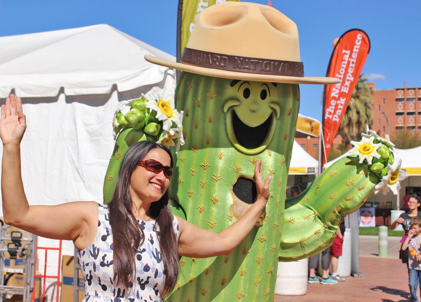 Woman posing with a large smiling cactus statue wearing a hat at an outdoor event.