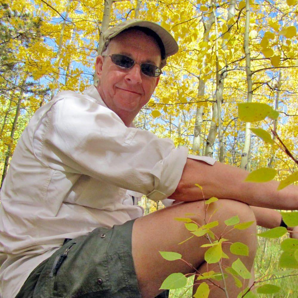 Man in sunglasses and cap sitting among yellow-leaved trees, smiling.