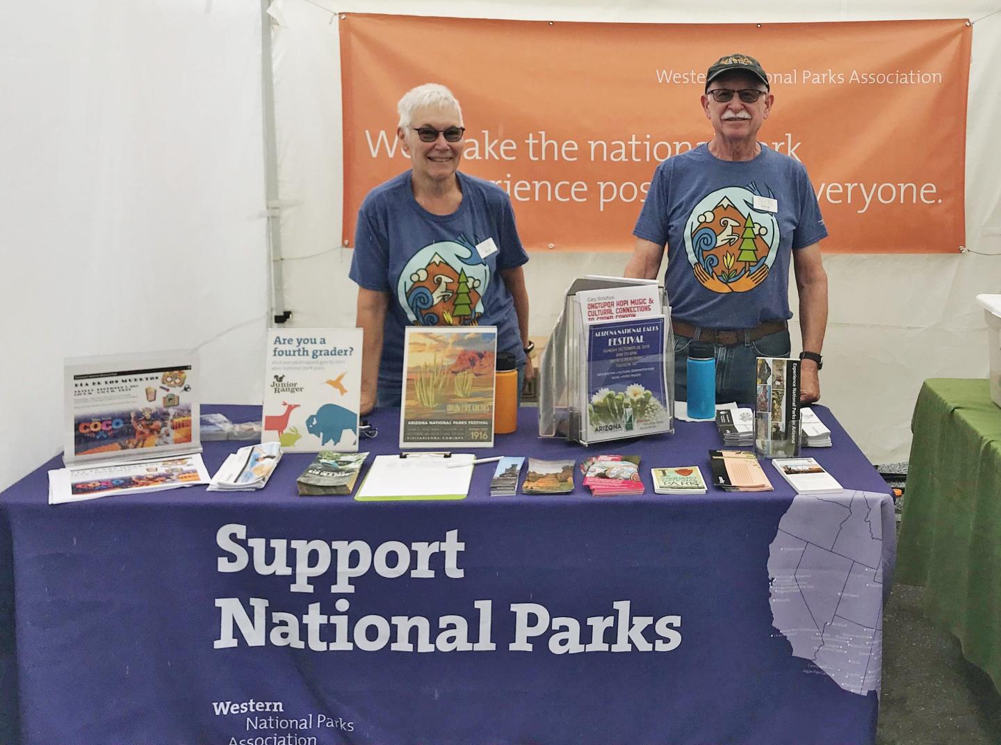 Two people stand behind a "Support National Parks" booth with brochures and posters.