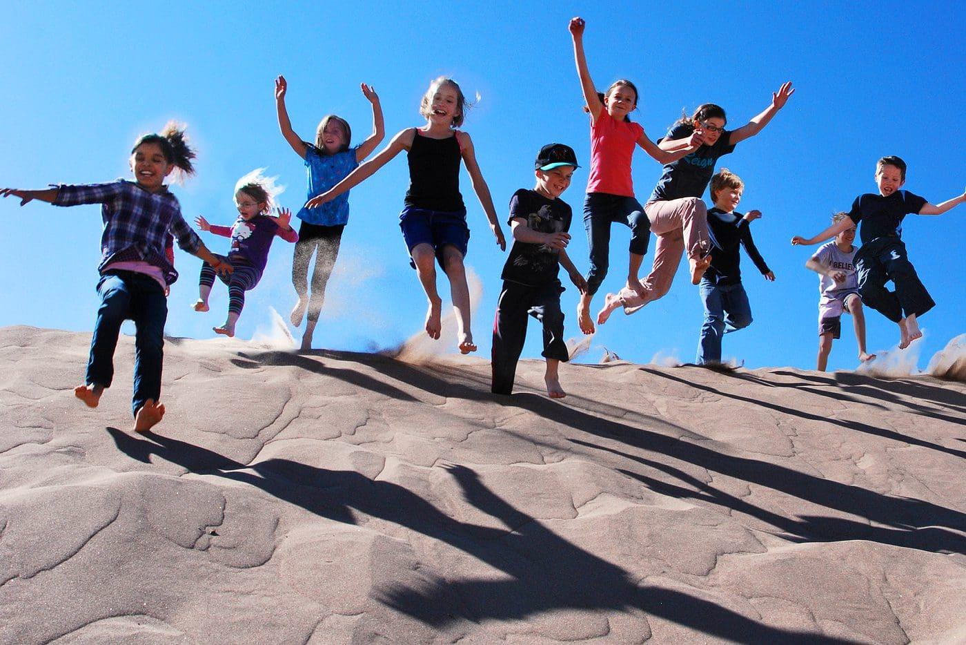 Children joyfully jumping on sand dunes under a clear blue sky.
