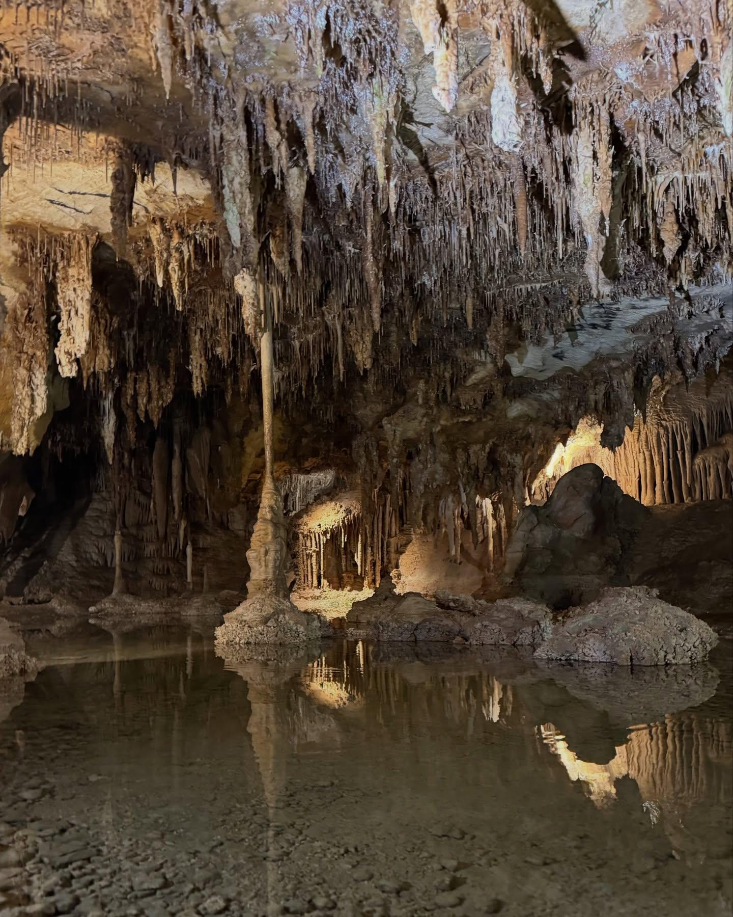Stalactites and stalagmites in a dimly lit cave with reflections in still water.