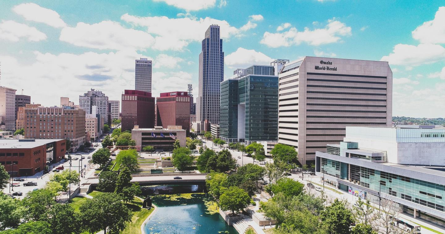 Omaha city skyline with tall buildings and a reflective pond beneath a bright blue sky.