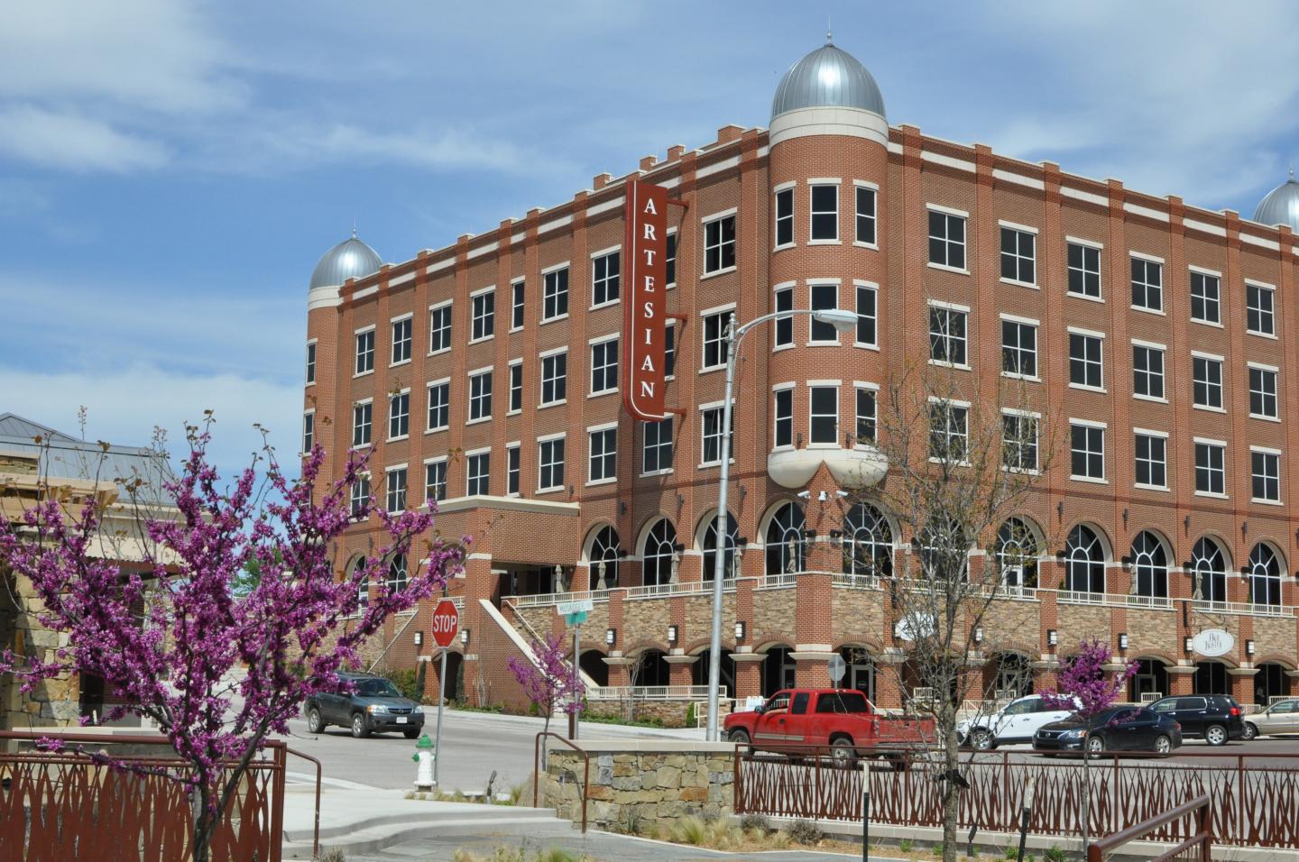 Artesian Hotel building with turret roofs, street view with purple blossoms and a red vehicle.