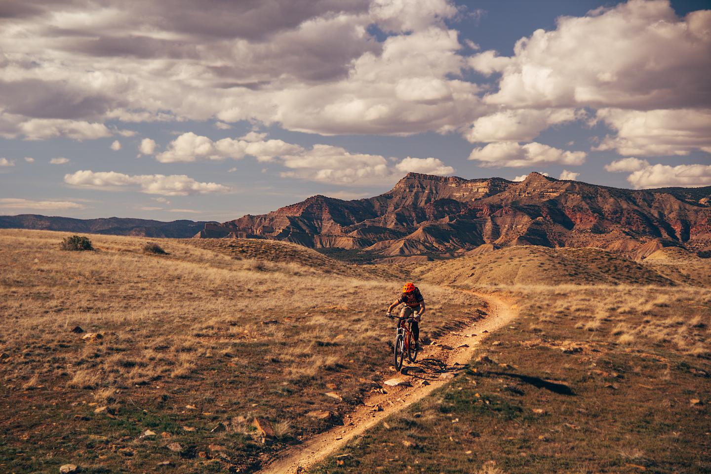 Cyclist on trail through dry landscape, mountains in background, under cloudy sky.