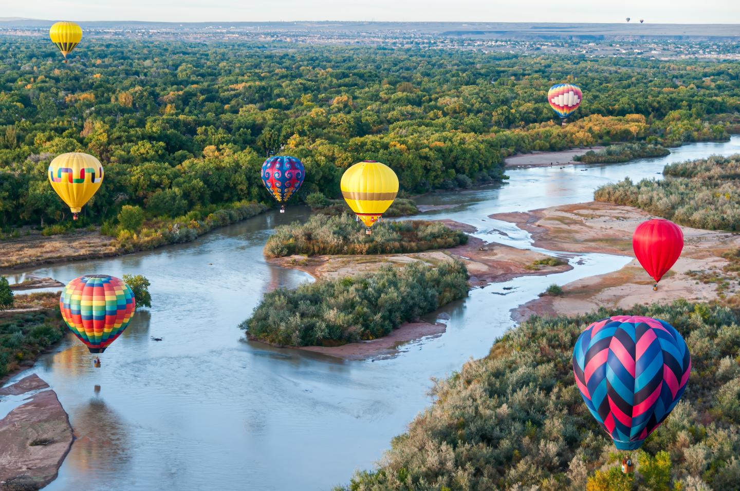Colorful hot air balloons float above a winding river and green landscape.