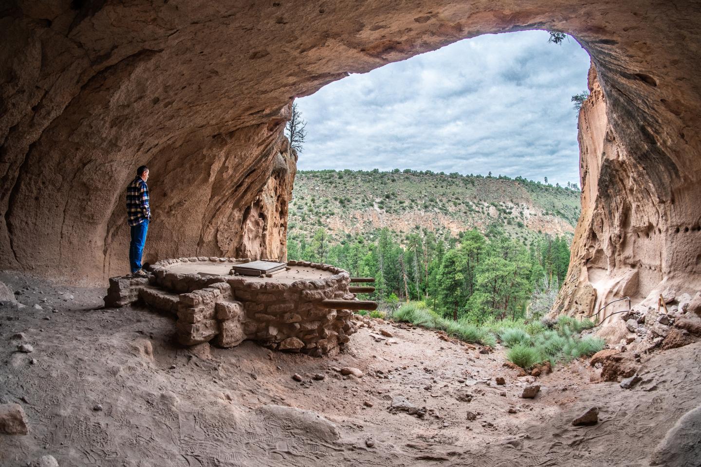 Alcove house opening overlooking forest, person standing near stone ruins.