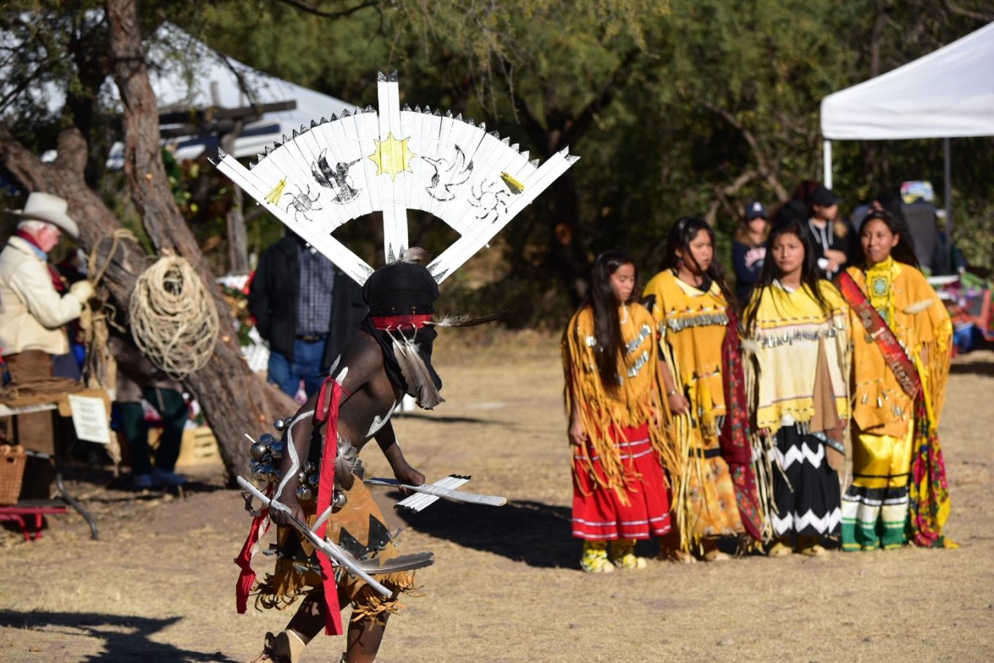 Dancer in traditional attire performs before four women in colorful dresses outdoors.