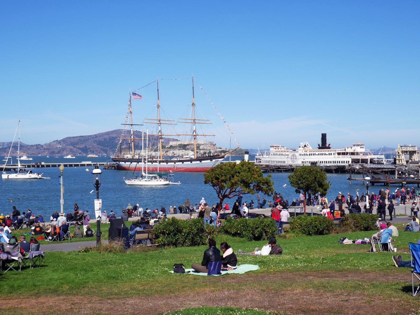 People relaxing in a park near San Francisco waterfront; a tall ship and ferry are in the background.
