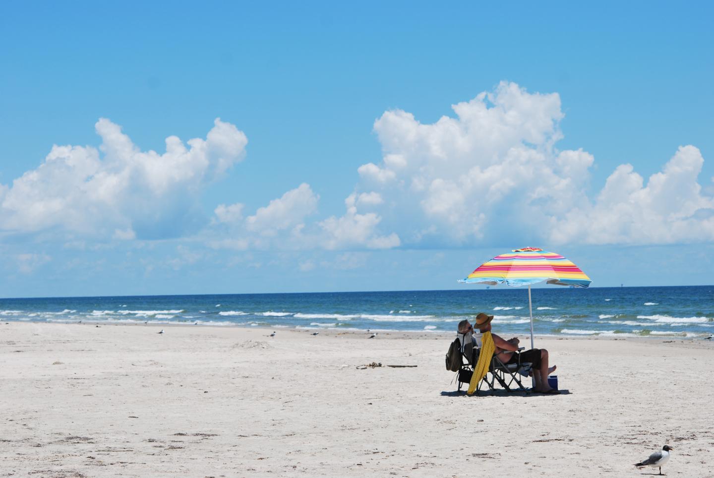 Sunny beach with people under a colorful umbrella, blue sky and ocean in the background.