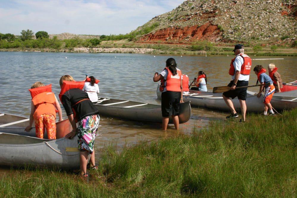 People in life vests preparing canoes by a lakeshore, grassy area, and red hills in the background.