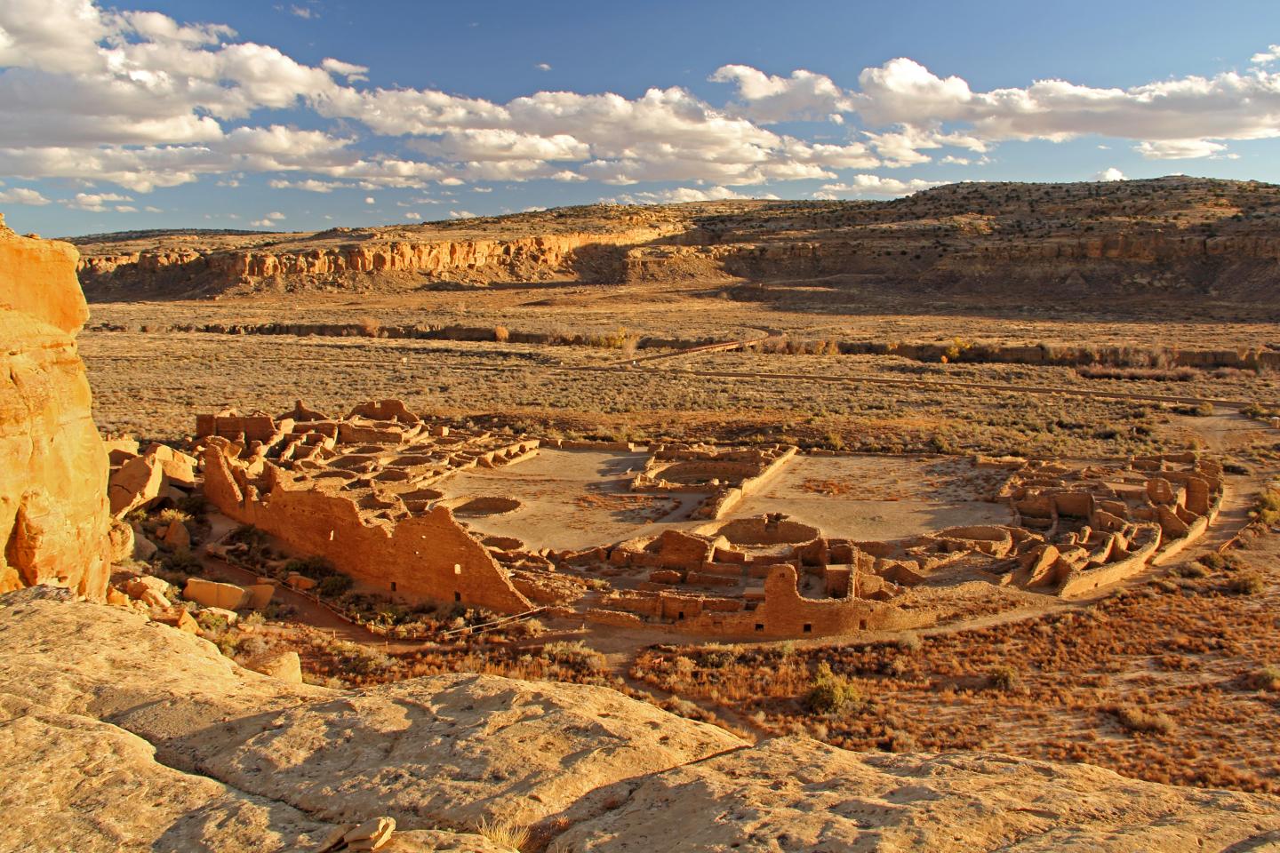 Chaco Culture ruins in a desert landscape under a partly cloudy sky.