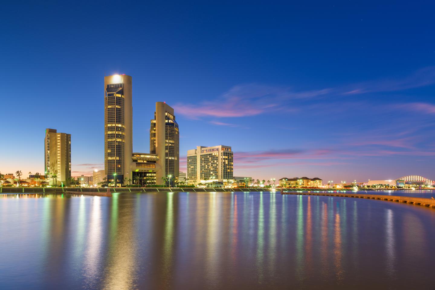 Corpus Christi city skyline reflecting on calm water at dusk with a vibrant blue sky.
