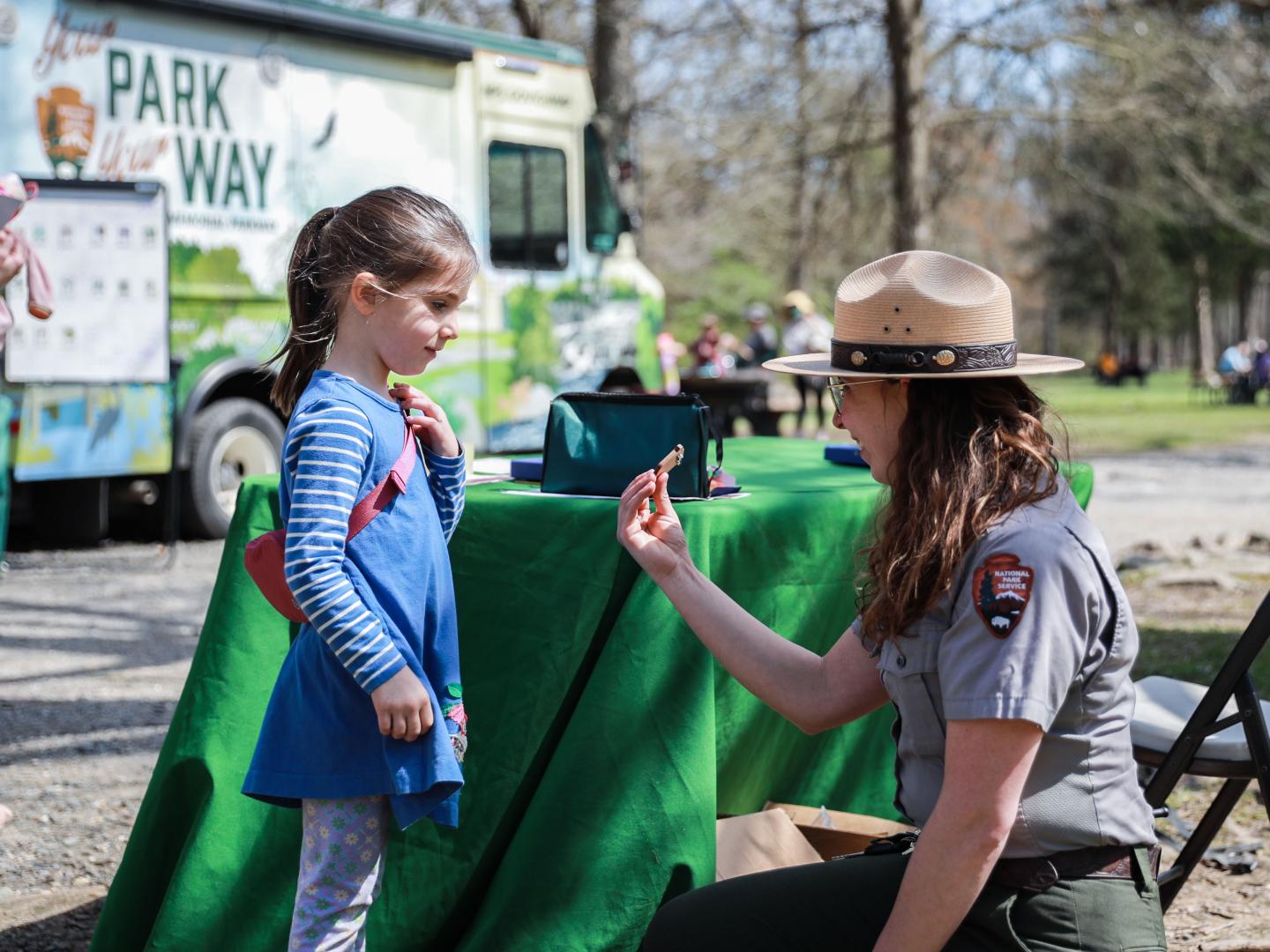 Park ranger kneeling, talking to a young girl in a park setting.
