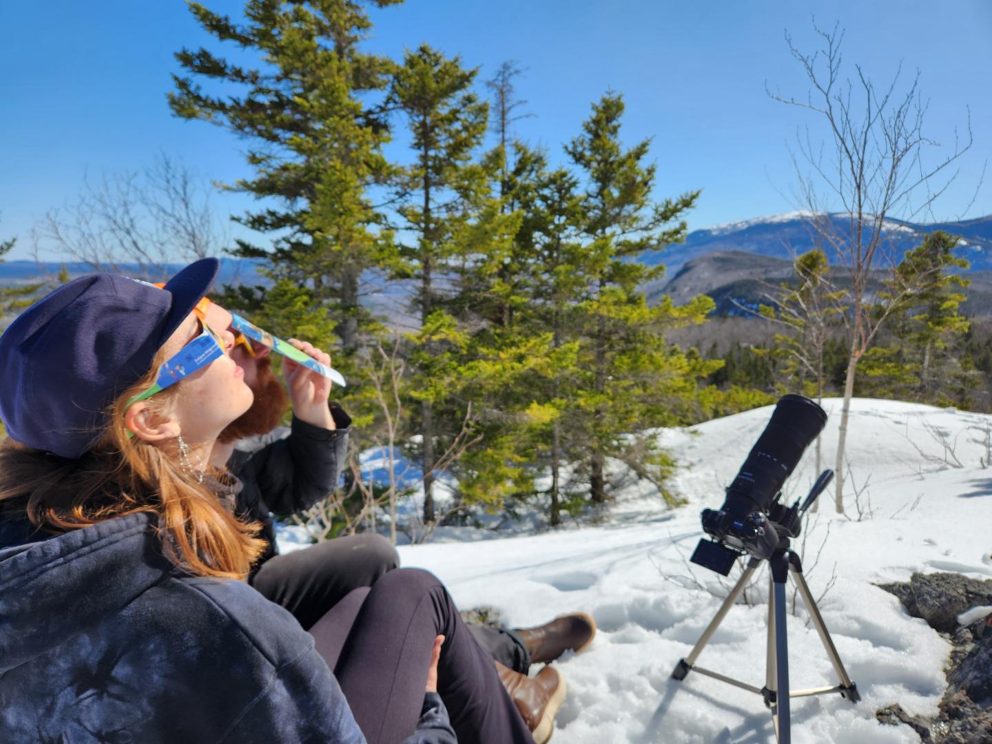 Woman viewing a solar eclipse through glasses, snowy mountain backdrop.