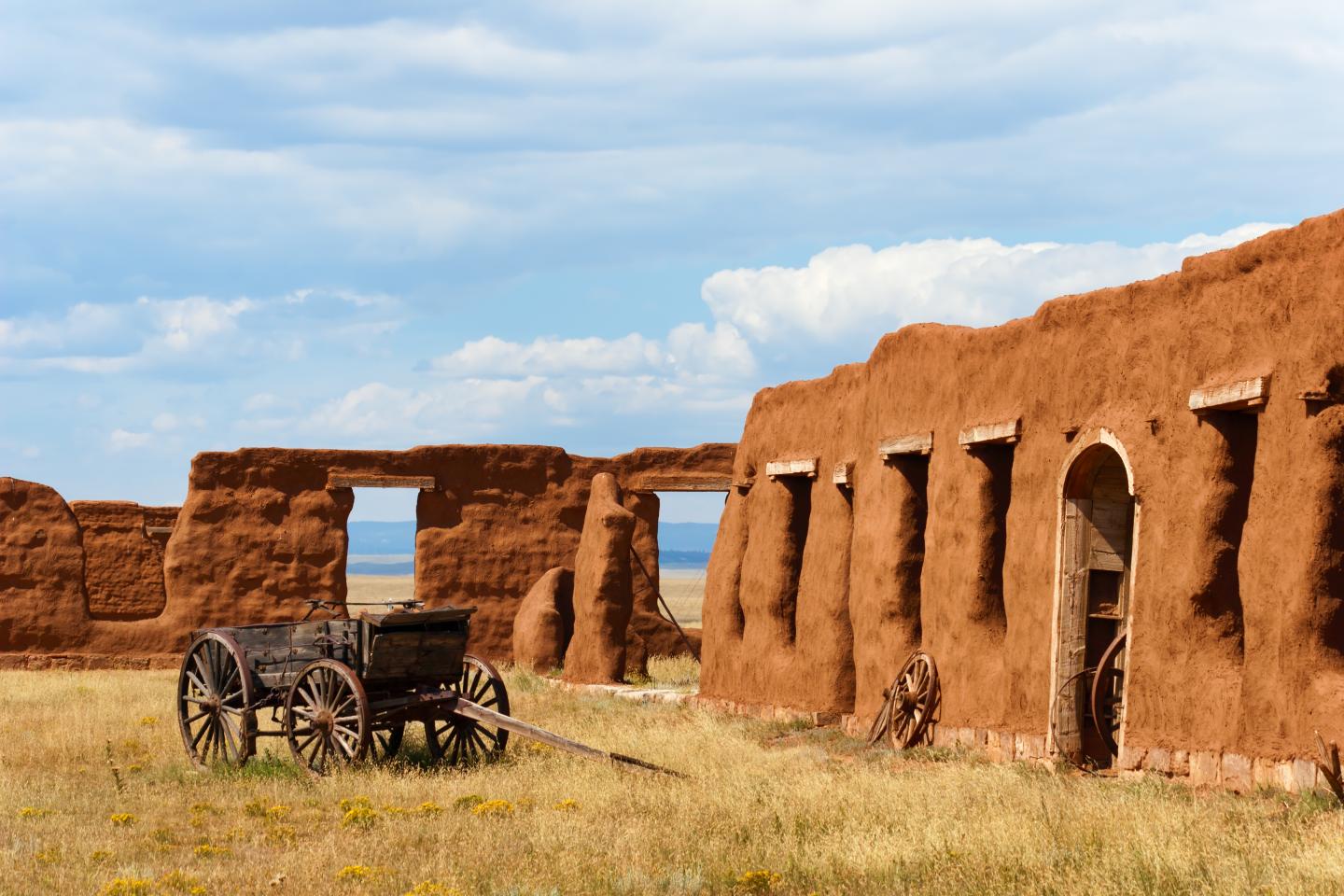 Old adobe structures and a wooden cart under a blue sky.