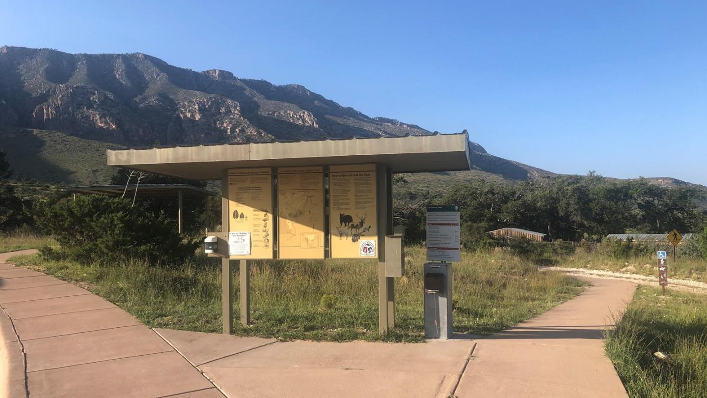 Trailhead signboard with mountains and clear sky in the background.