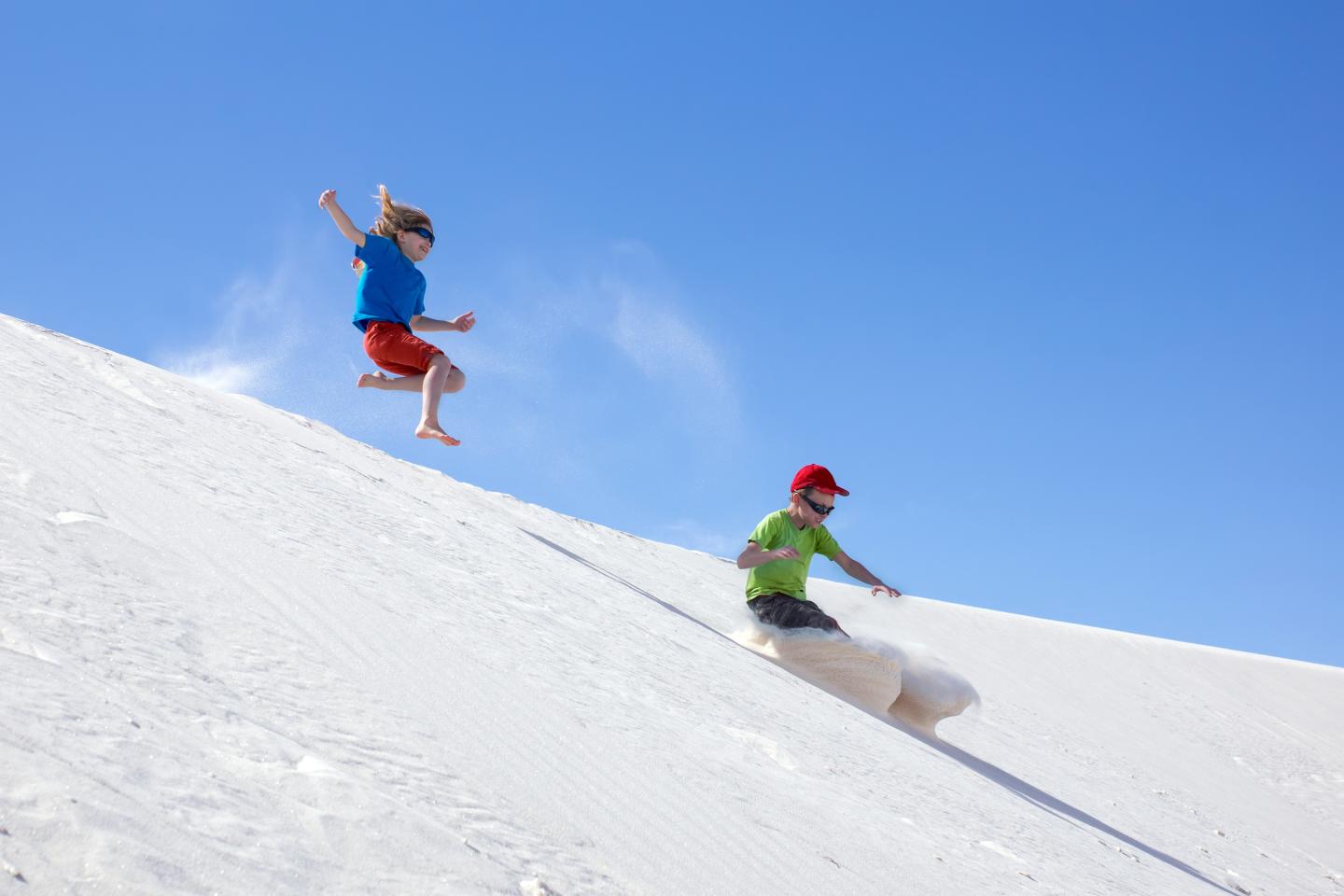 Kids joyfully sliding down a sand dune under a clear blue sky.