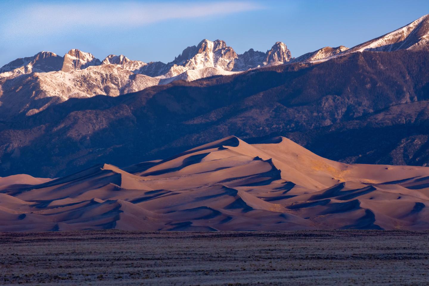 Sand dunes with snowy mountains in the background at sunset.