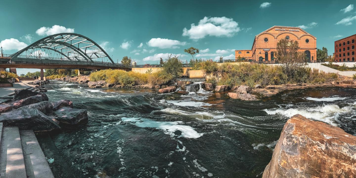River with rushing water, arched bridge, and historic buildings under a clear sky.
