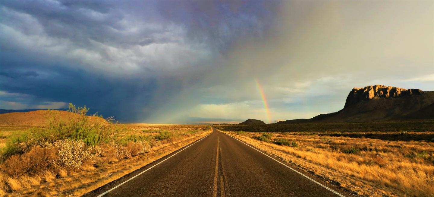 Open road with rainbow, dramatic clouds, desert landscape, and distant rock formation.