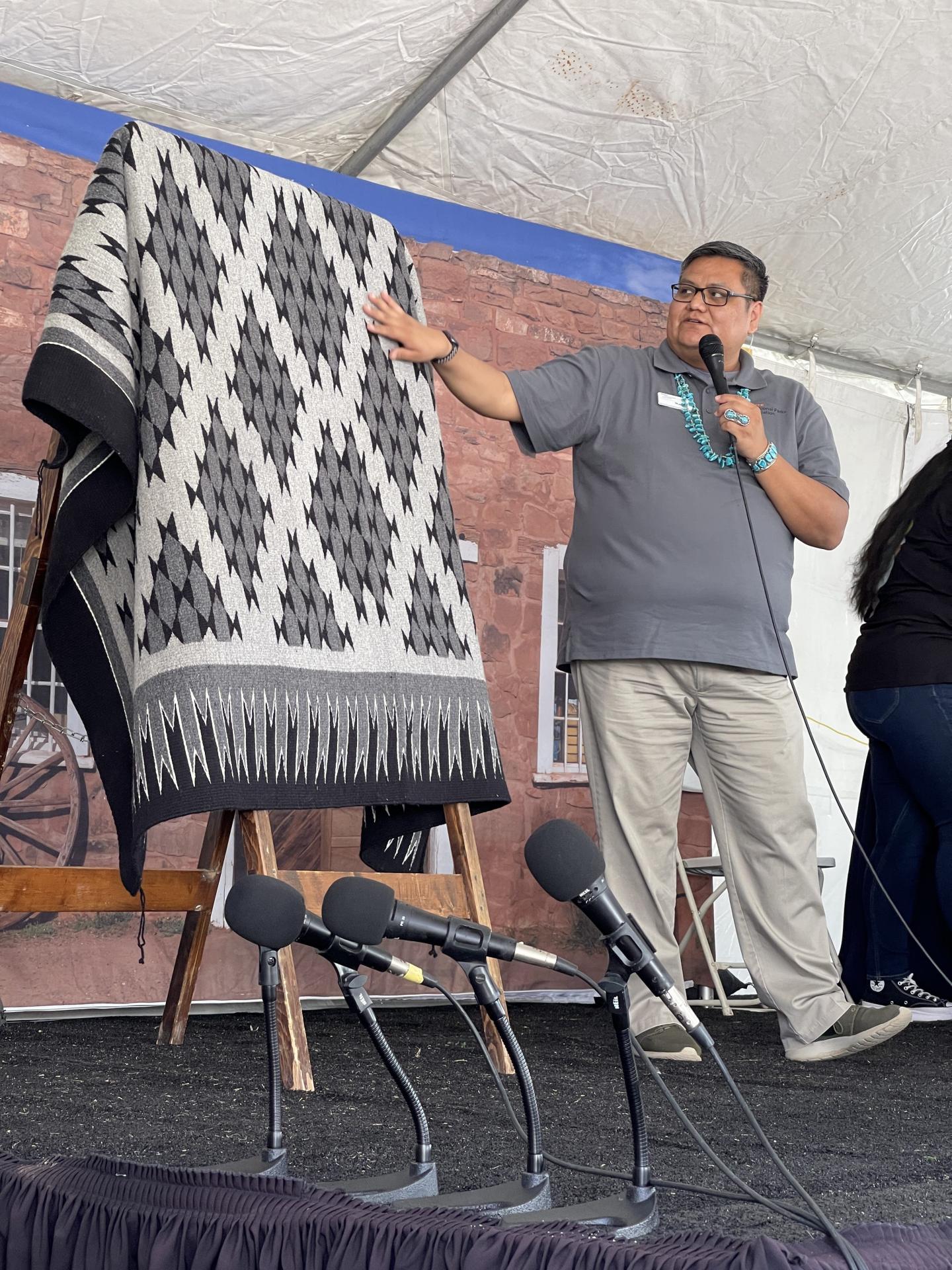 Man speaking into a microphone, showing a patterned textile on stage.