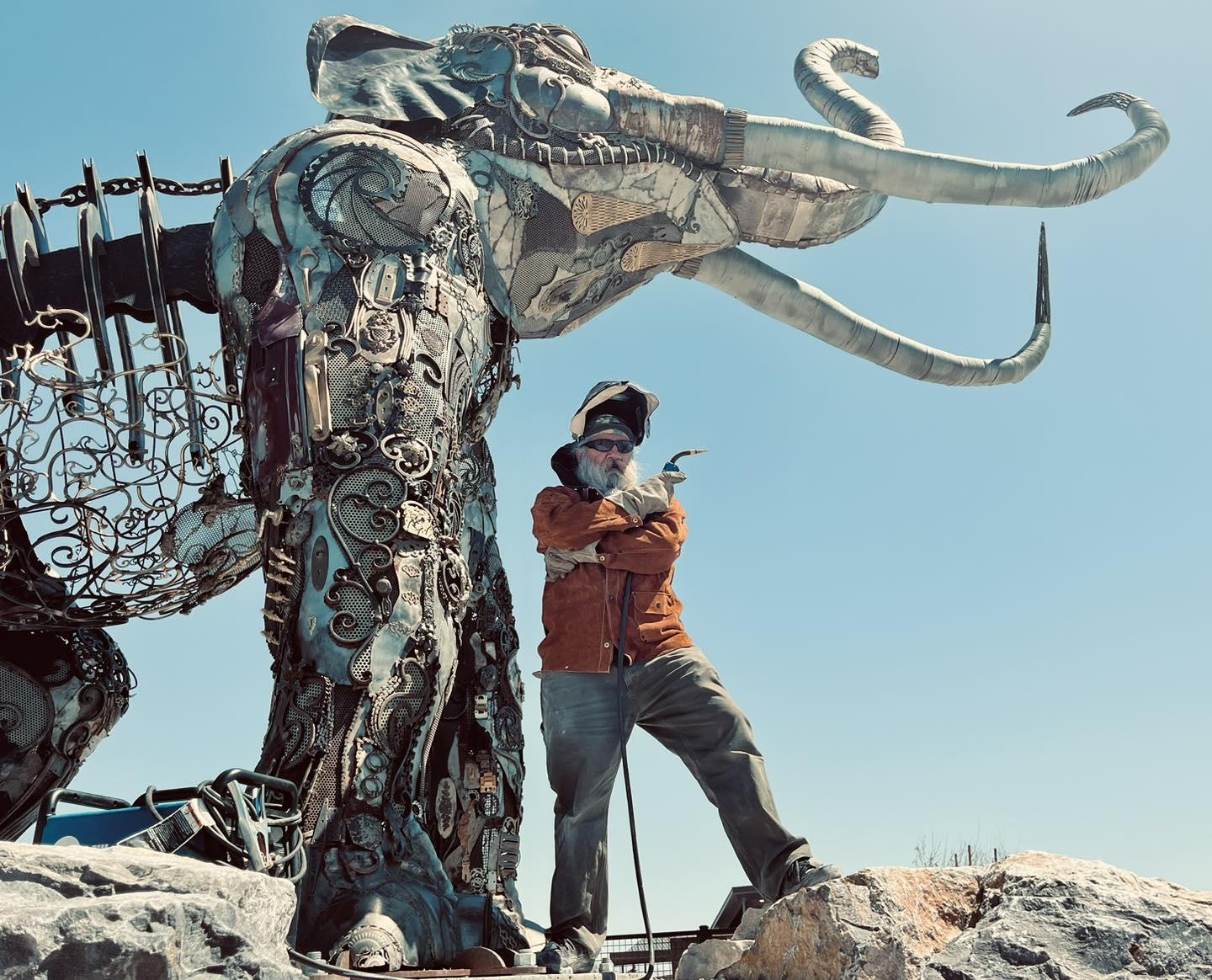 Person posing with large metal mammoth sculpture under blue sky.