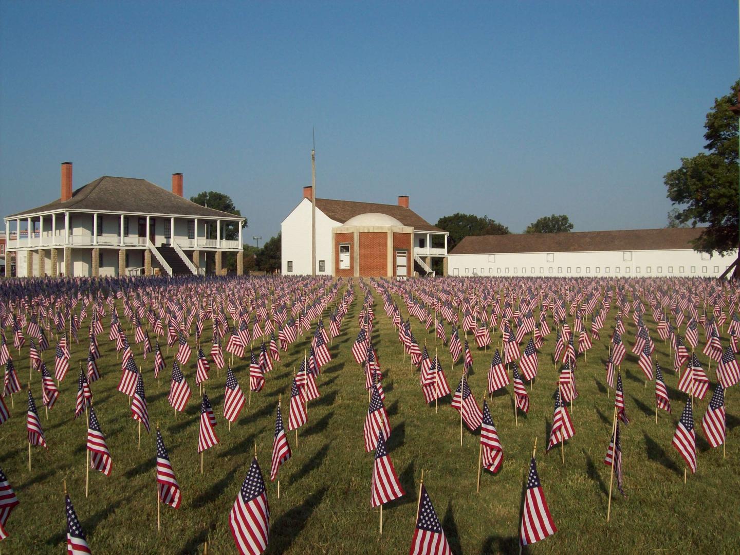 Field of American flags in front of infantry barracks at Fort Scott under a clear blue sky.