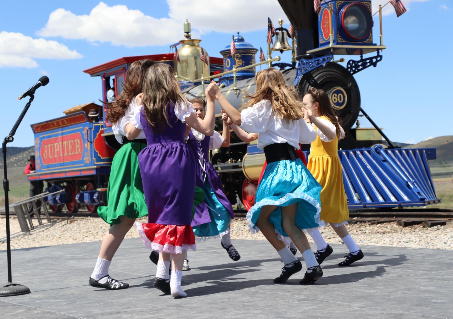 Children in colorful dresses dancing near a bright vintage train.