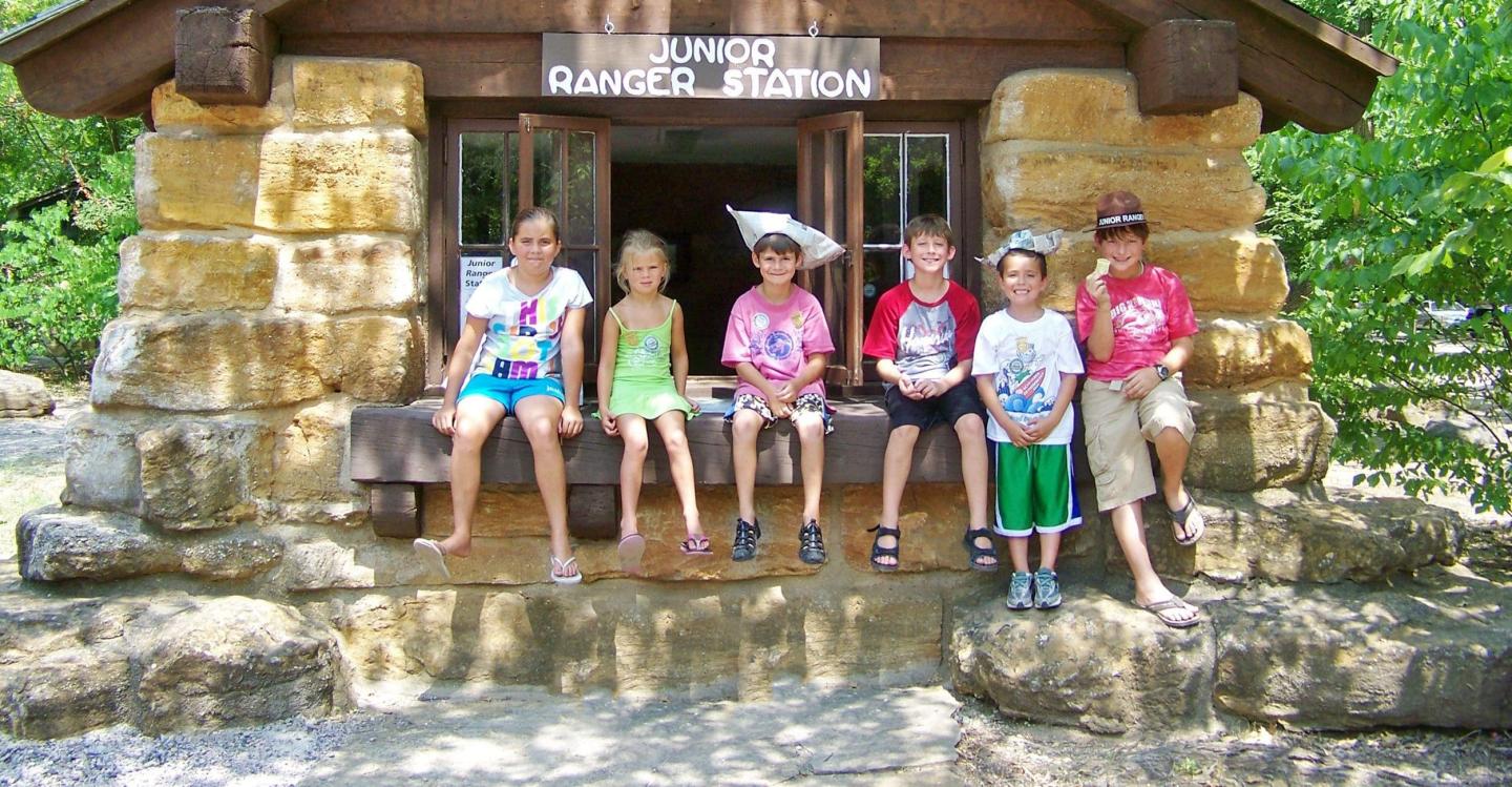 Children sitting on a large rock outside a rustic cabin in sunlight.