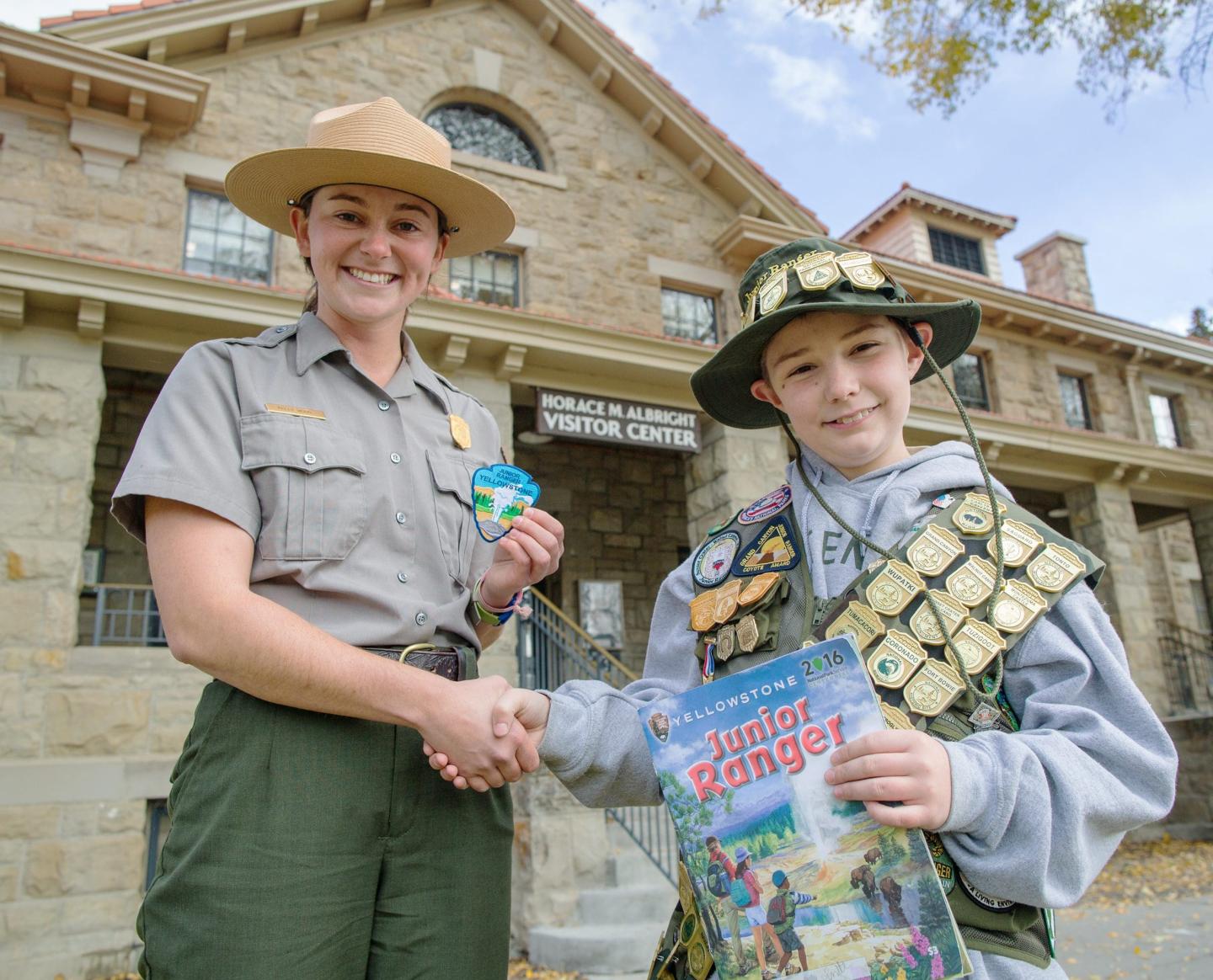 Park ranger and child in uniform smiling, shaking hands outside a stone building.