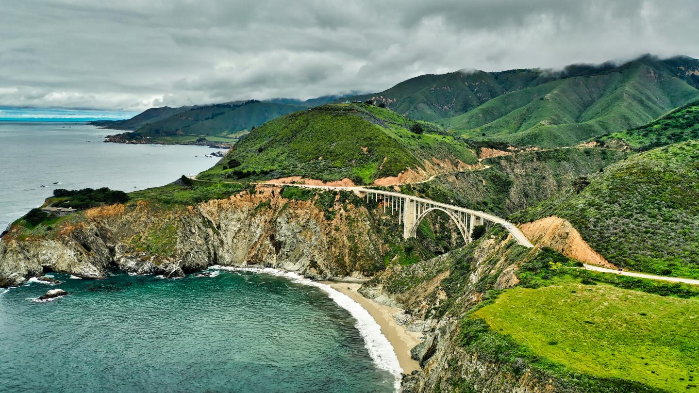 Coastal cliff with a curving road and green hills under a cloudy sky.