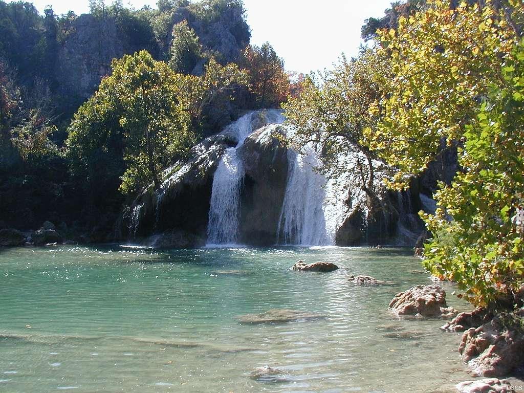 Turner Falls flowing into a serene, green pool surrounded by trees.
