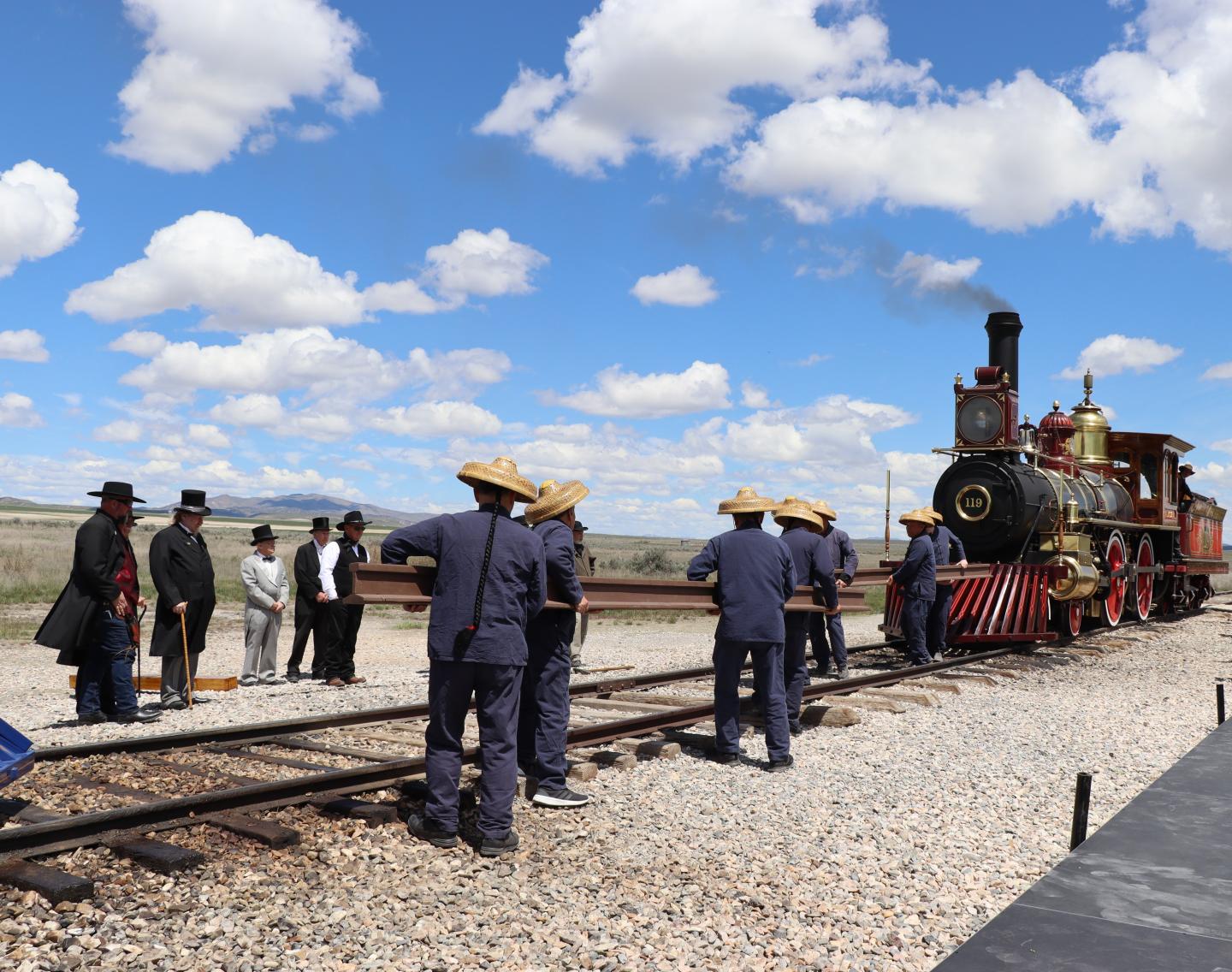 Historic steam locomotive with people in period costumes by tracks under blue sky.