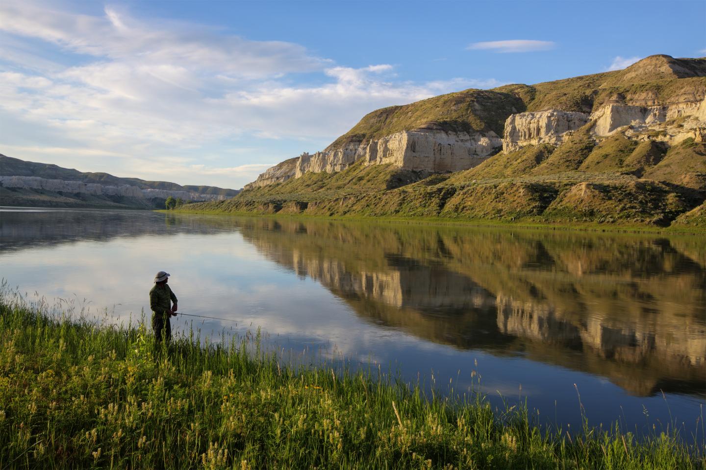Person stands by a calm lake with distant cliffs and a clear sky.