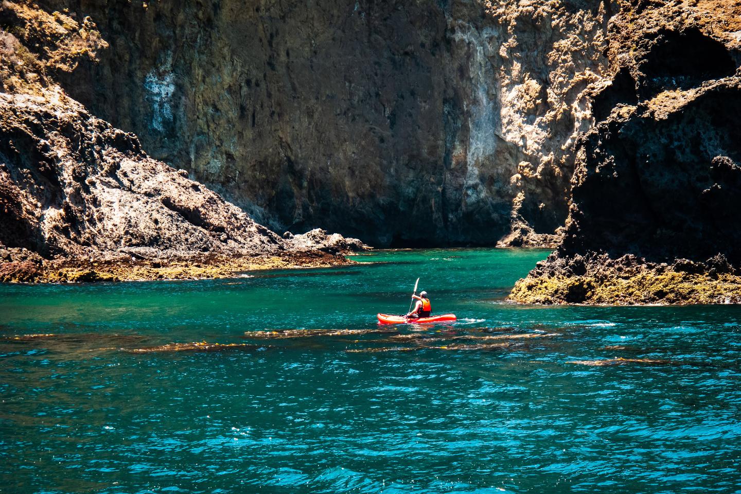 Kayaker in red boat on clear blue water between cliffs.