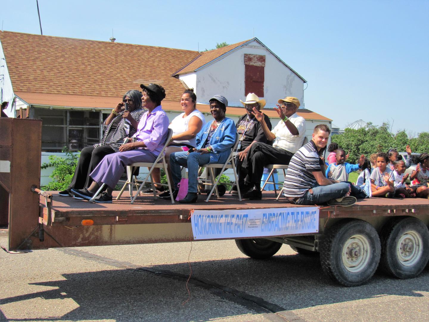 People seated on chairs on a parade float in sunny outdoor setting at Nicodemus Homecoming celebration..
