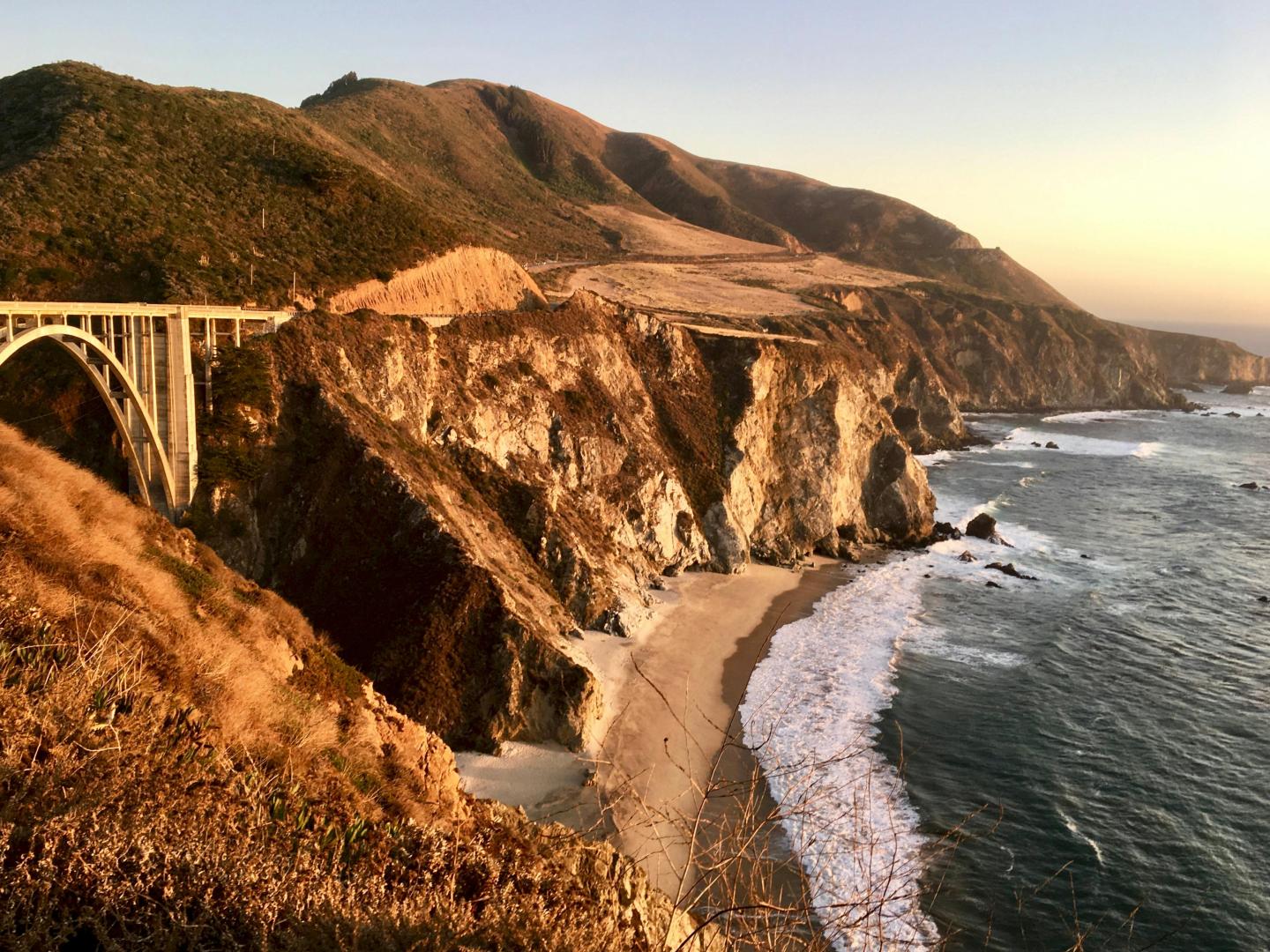 Coastal cliffs at sunset, ocean waves hitting sandy shore.