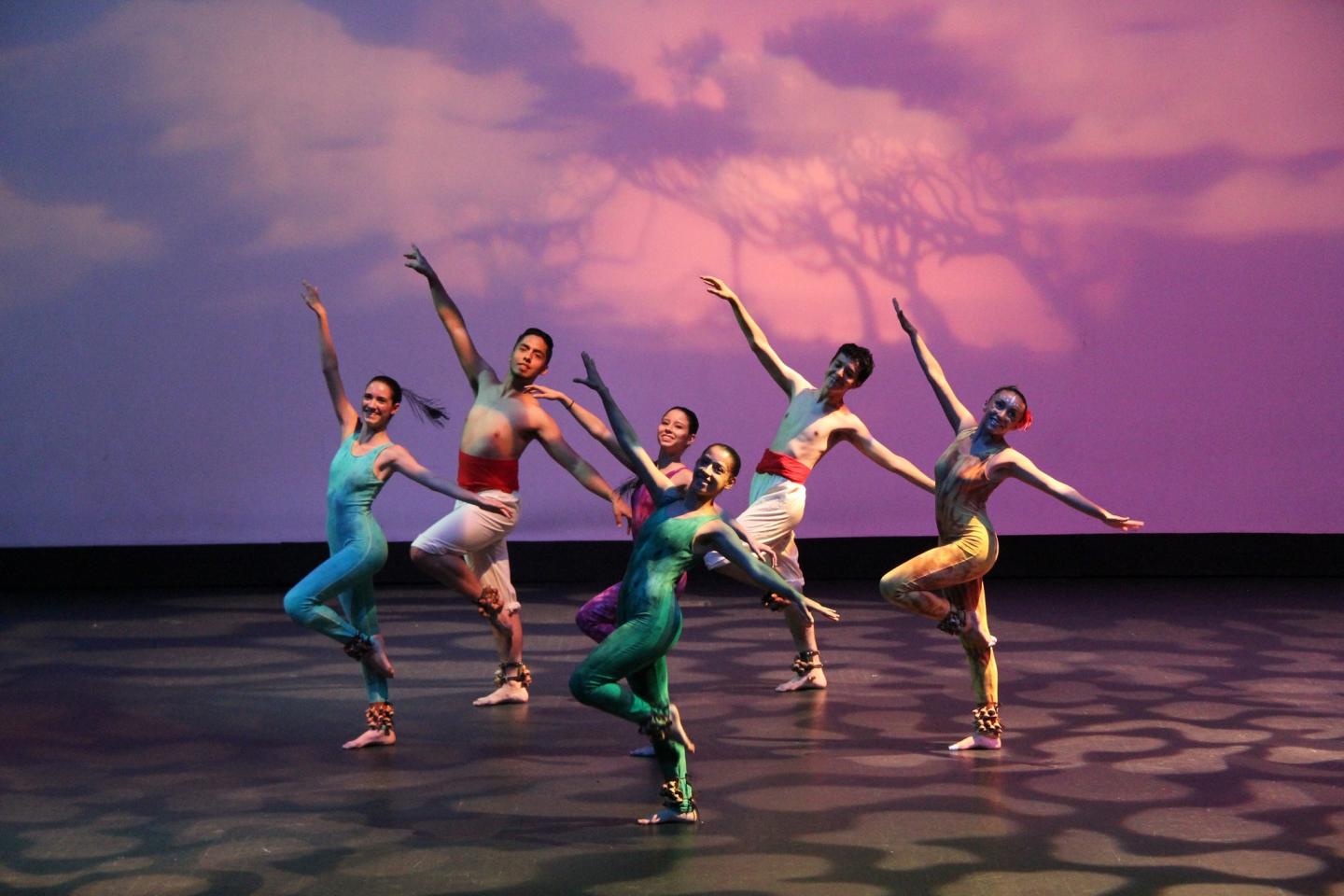 Six dancers in colorful costumes perform on stage with a purple backdrop.