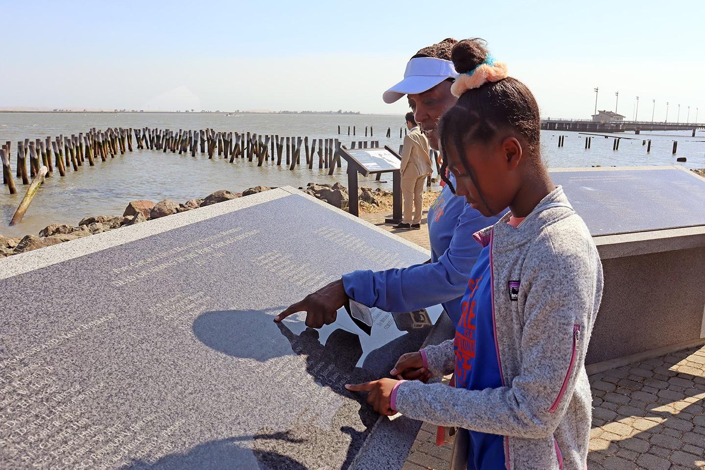 Two people read a text panel outdoors near a water view at a memorial at Port Chicago Naval Magazine National Memorial.