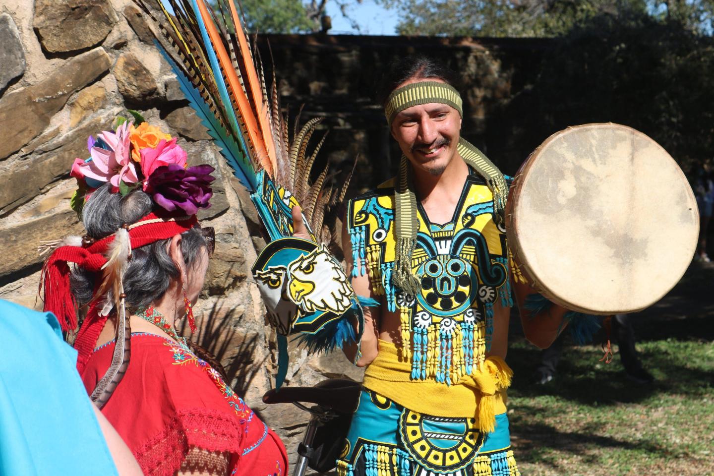 Man in colorful traditional attire holding a drum outdoors.
