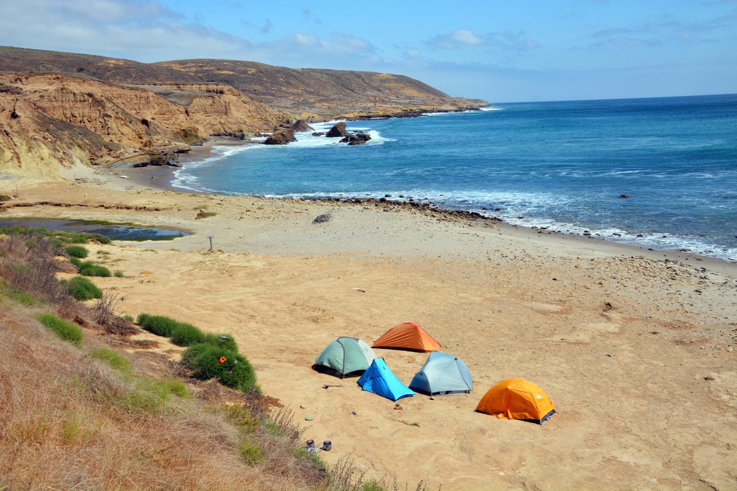 Tents on a sandy beach by blue ocean, with cliffs in the background.