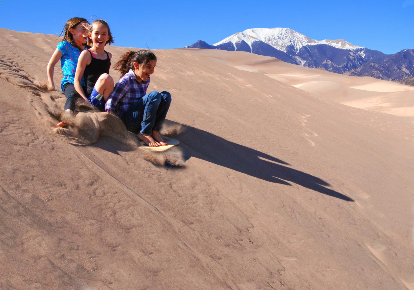 Three kids sliding down a sand dune, mountains in the background, bright blue sky.